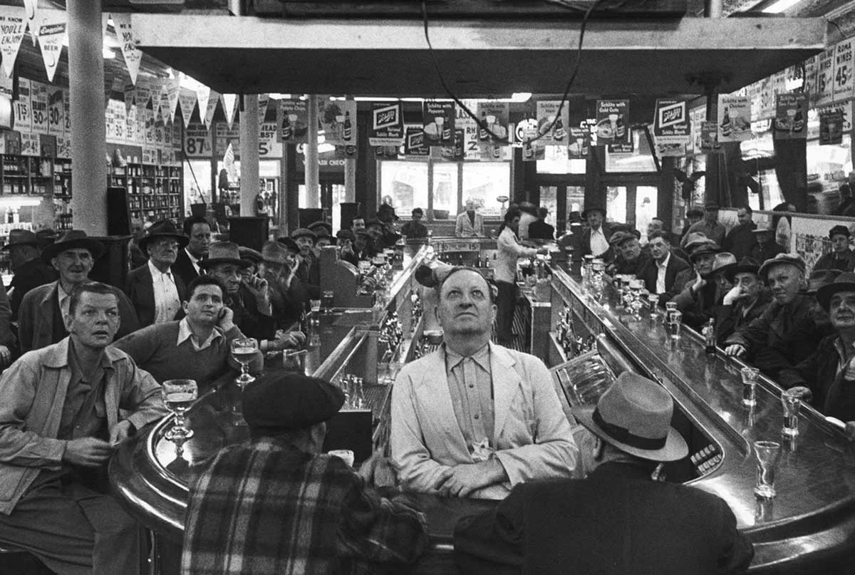 A group of people, mostly men, sit and stand around a bar, watching something intently. Glasses and posters fill the lively, busy space. The scene is in black and white.