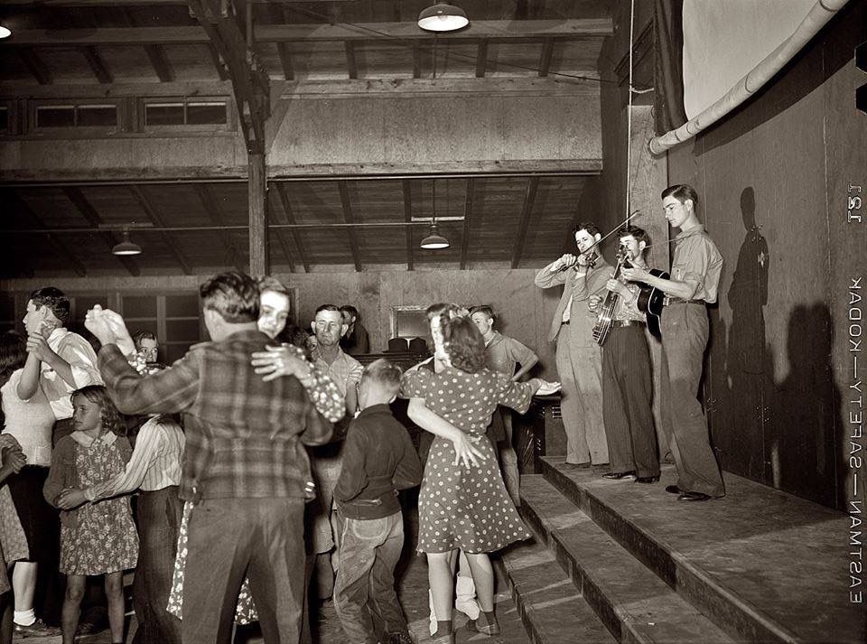 Couples dance in a hall while a small band plays on stage. The scene appears vintage, with people in patterned clothes and dim lighting. Musicians play violin, saxophone, and guitar as others enjoy dancing below.