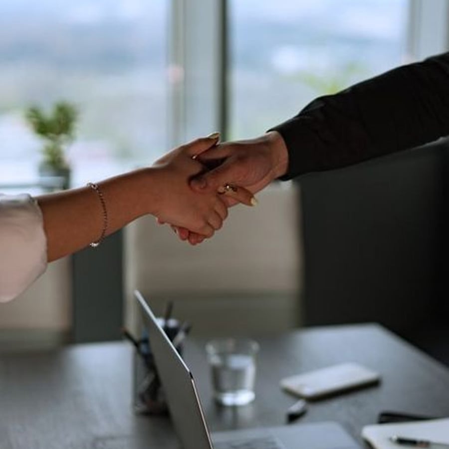 Two people shake hands across a desk with a laptop, phone, and glass of water on it. A window and blurred outdoor scenery are visible in the background.