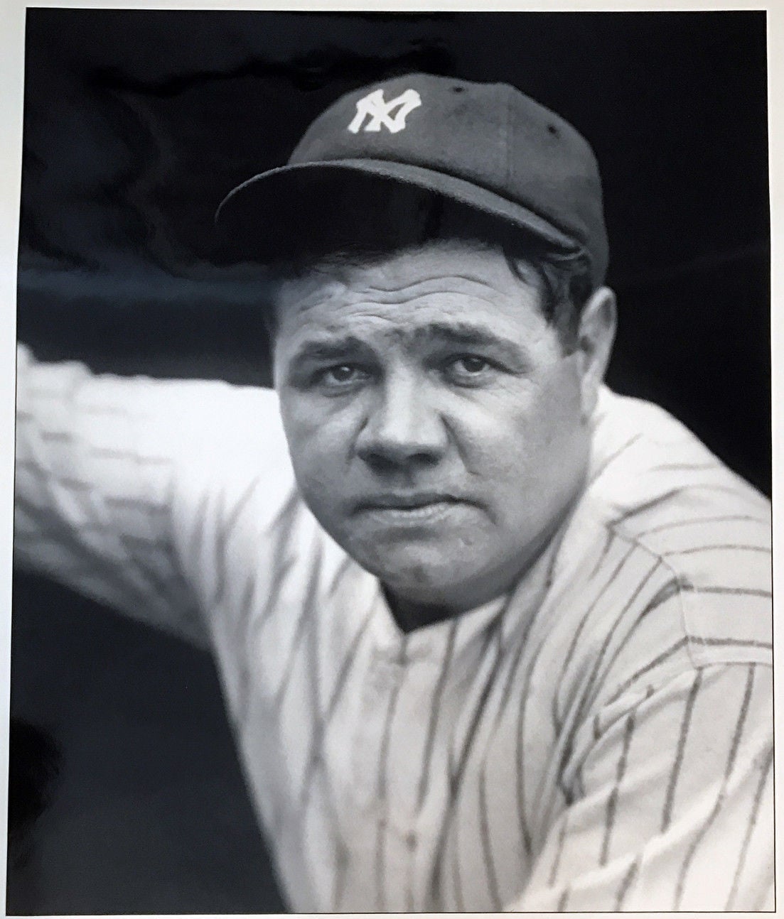 A baseball player in a pinstriped jersey and New York Yankees cap looks at the camera with a serious expression. The photo is black and white.