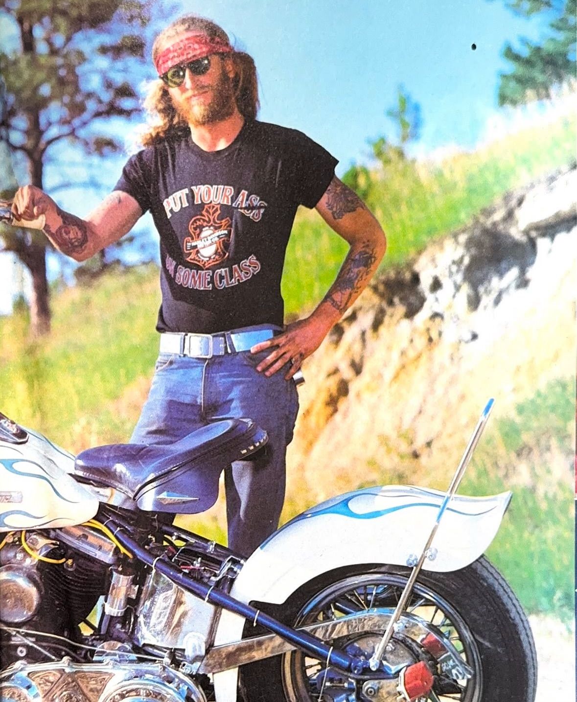 A bearded man in sunglasses and a bandana stands outdoors beside a custom motorcycle. He wears a black T-shirt with a graphic, blue jeans, and poses with one hand on his hip.