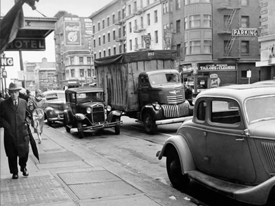 Black and white photo of a busy city street in the 1940s with vintage cars parked along the curb, people walking on the sidewalk, and a large delivery truck driving down the road.