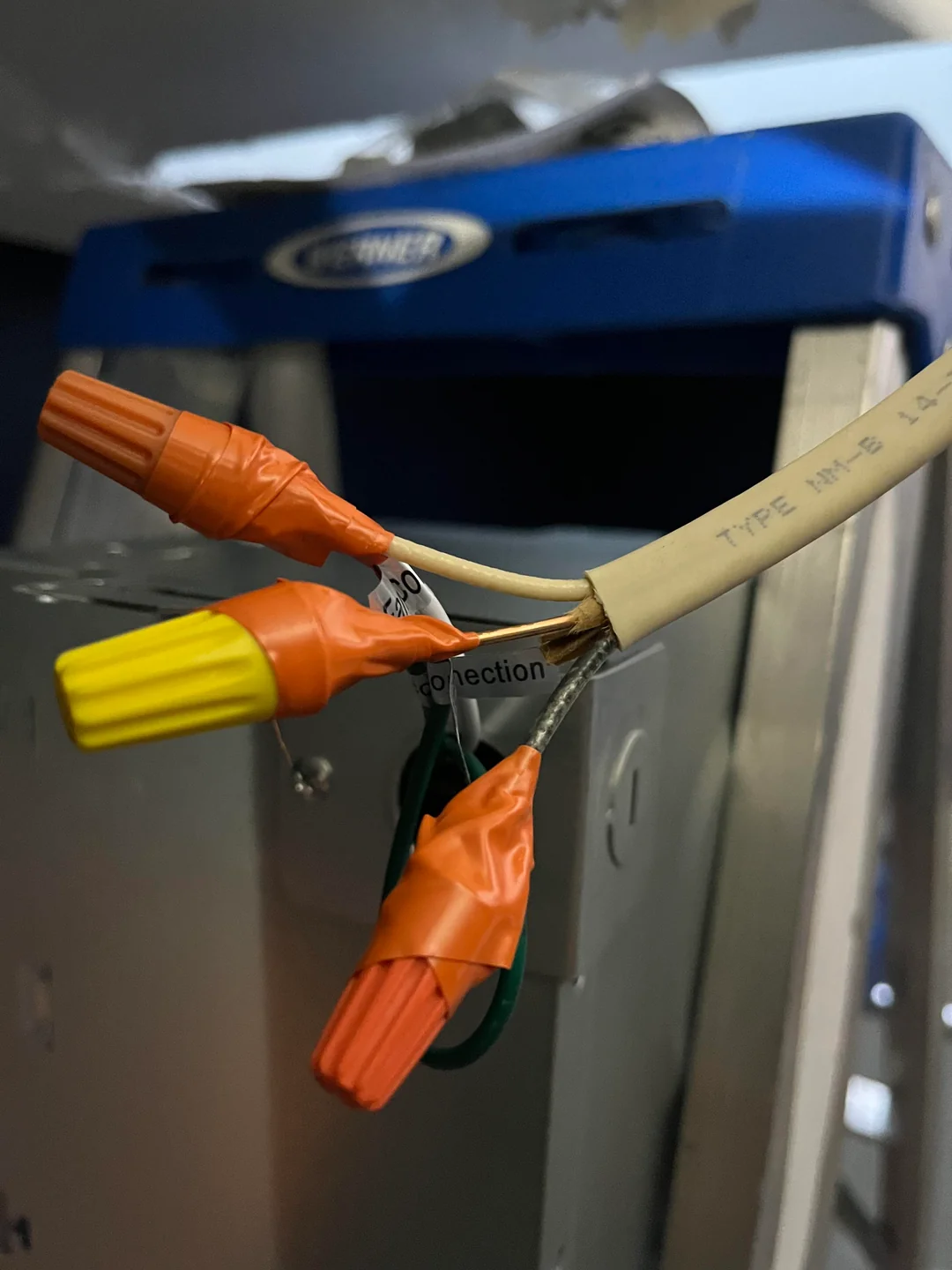 Close-up of electrical wires with orange and yellow wire nuts, wrapped in orange tape. The wires are exposed and hanging in front of a metal unit, with a blue Carrier-branded device in the background.