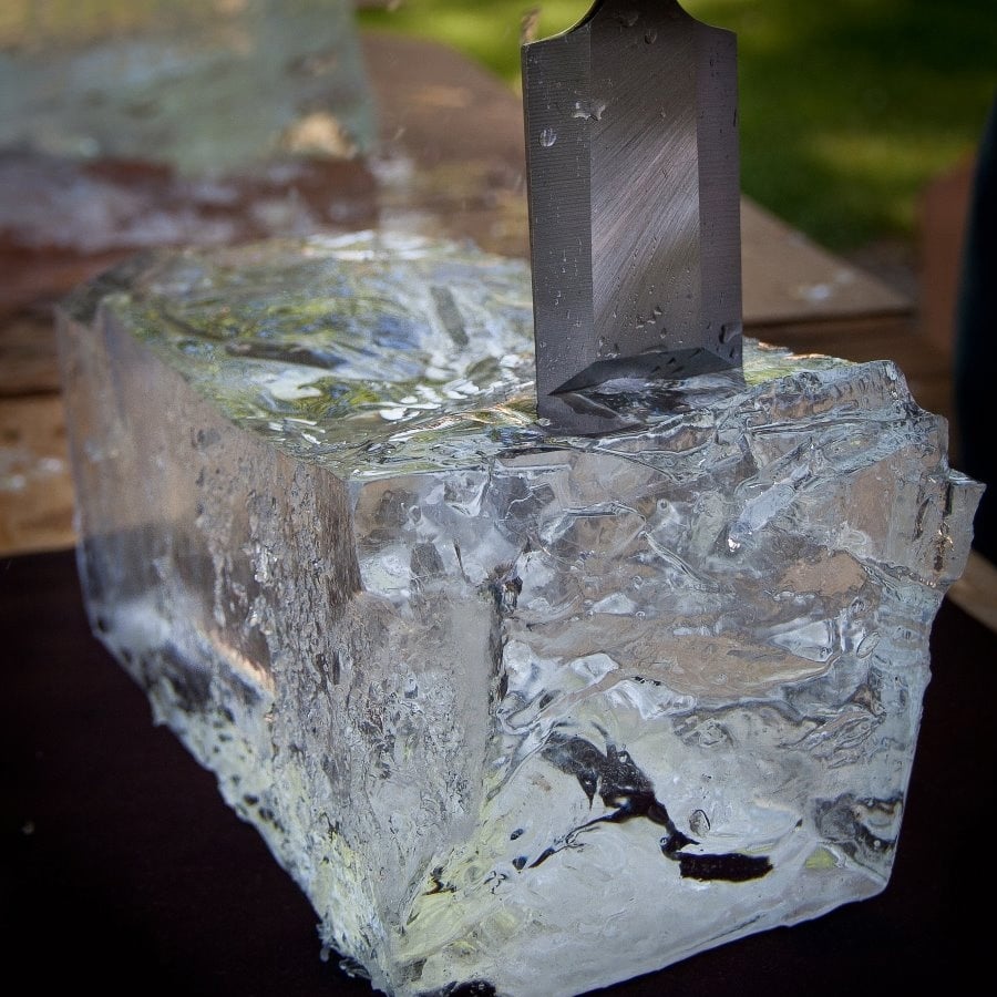A large knife is stuck into a clear rectangular block of ice resting on a dark surface, with water droplets visible on the ice.