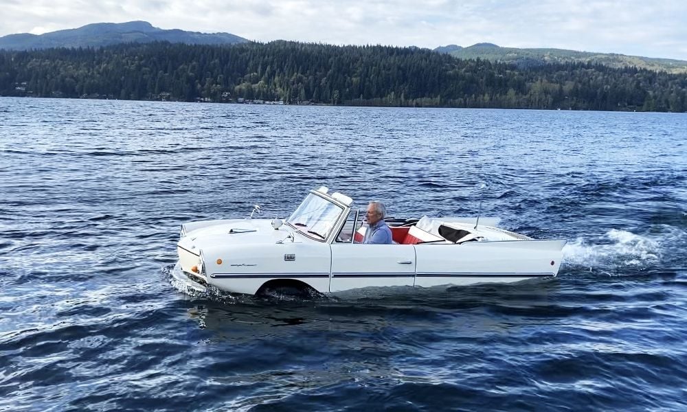 A man drives a white amphibious car across a lake, surrounded by forested hills and mountains under a partly cloudy sky.