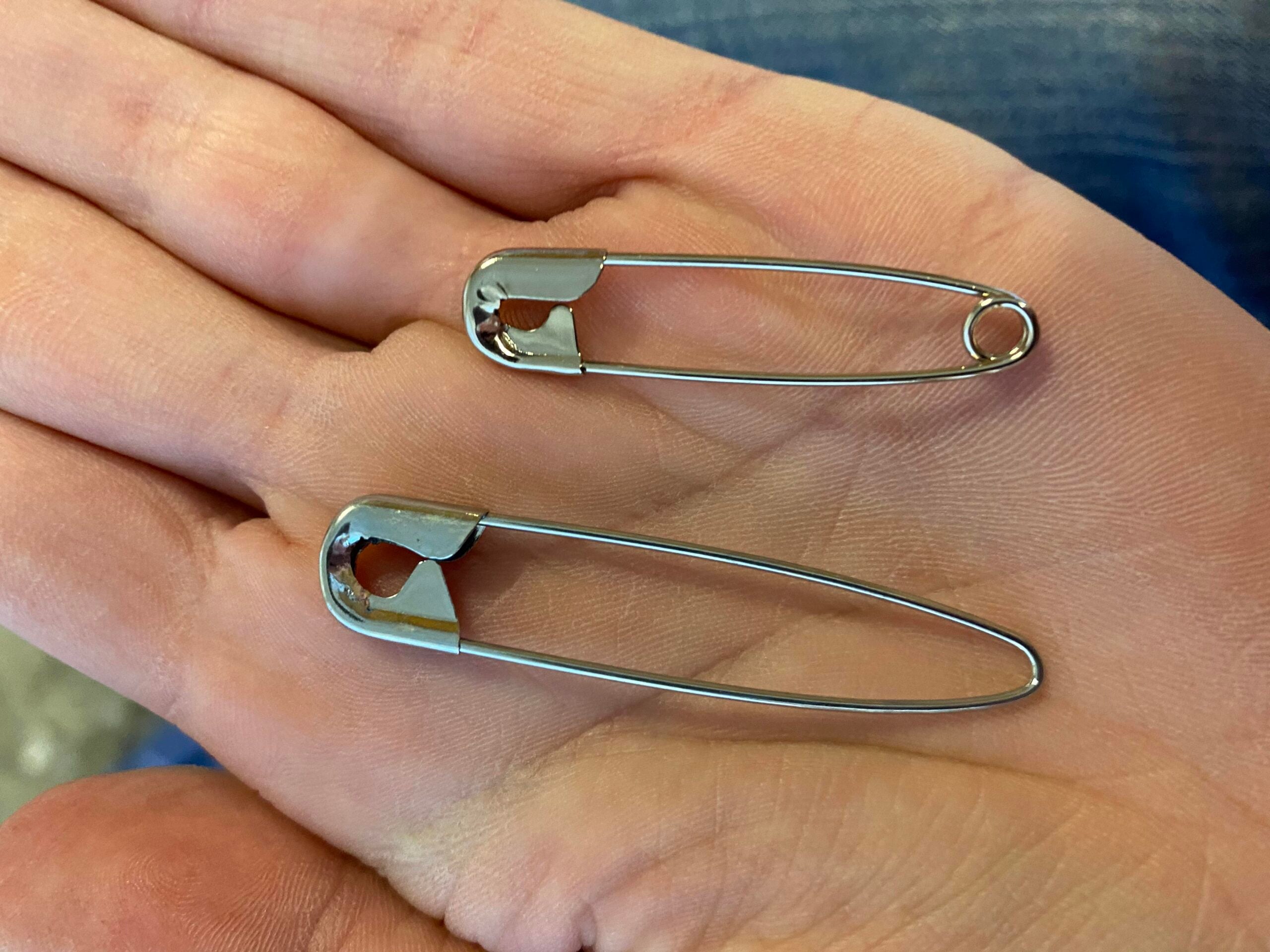 A hand holding two silver safety pins, one large and one small, with denim jeans visible in the background.