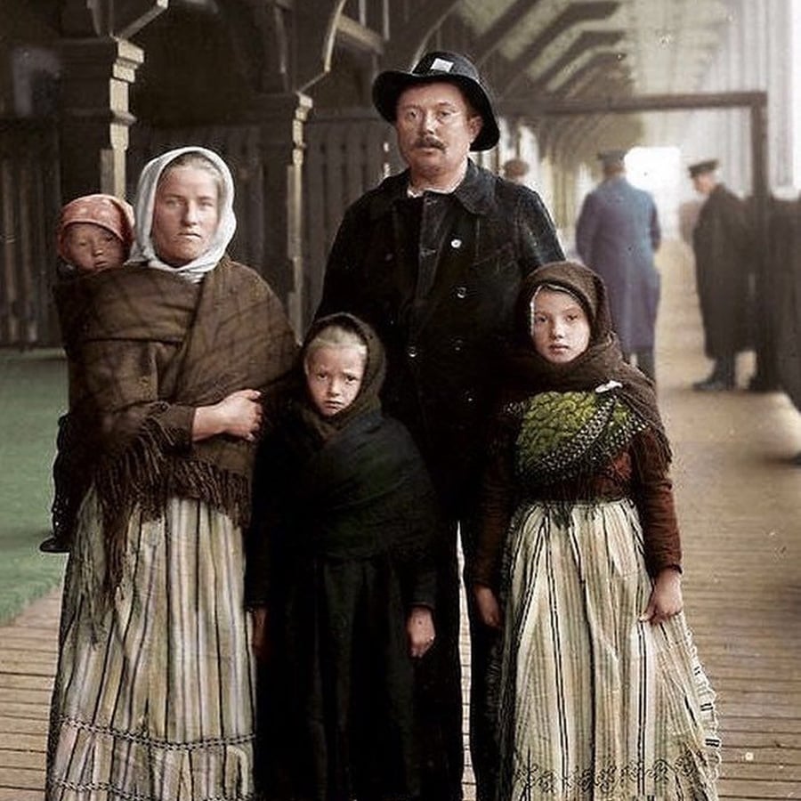 A family of five, dressed in old-fashioned clothing, stands together in a covered walkway. The woman holds a baby, while two girls stand in front. The man, wearing a hat, stands at the center.