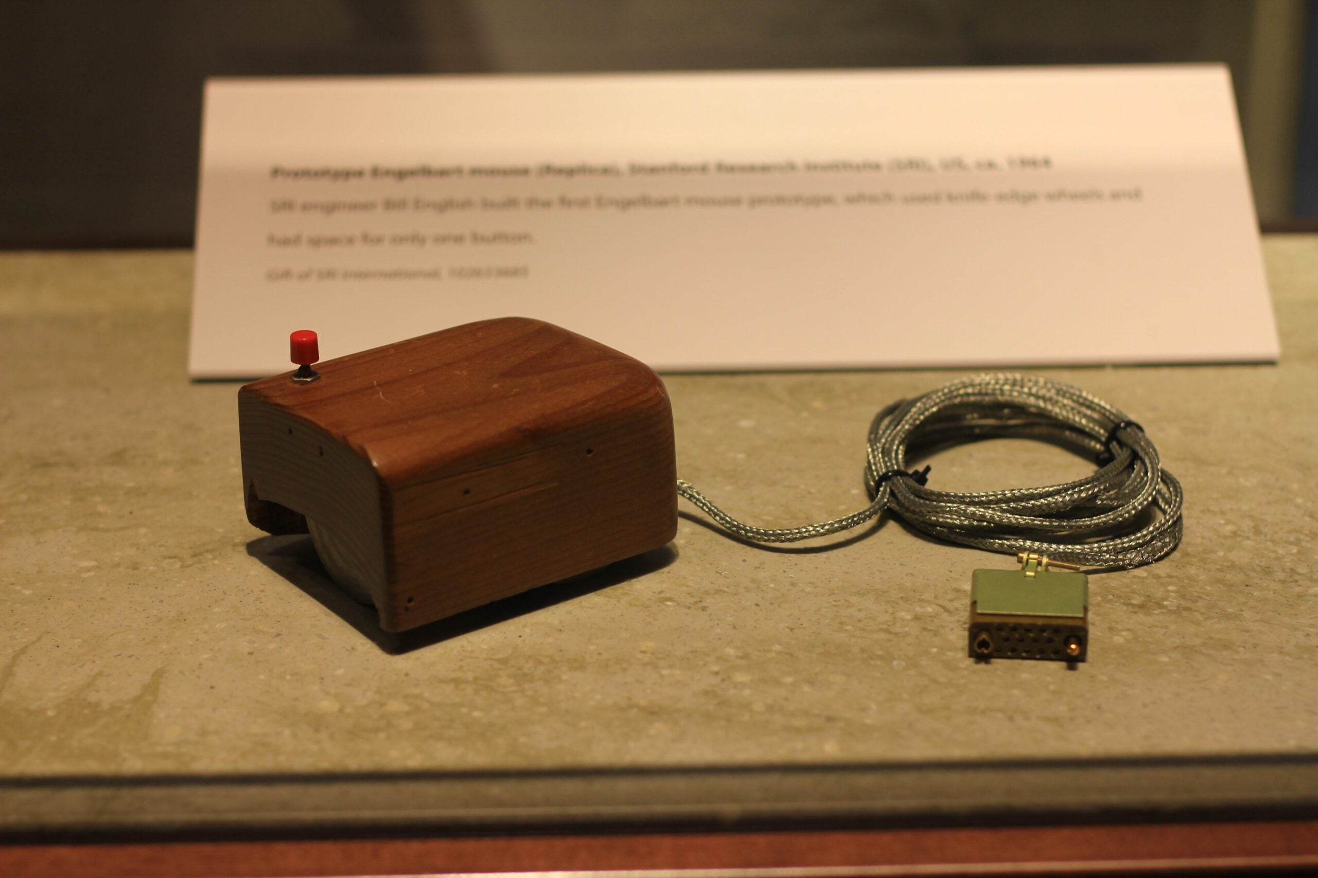 A wooden prototype computer mouse with a single red button, attached to a cord, displayed on a flat surface with an informational plaque in the background.