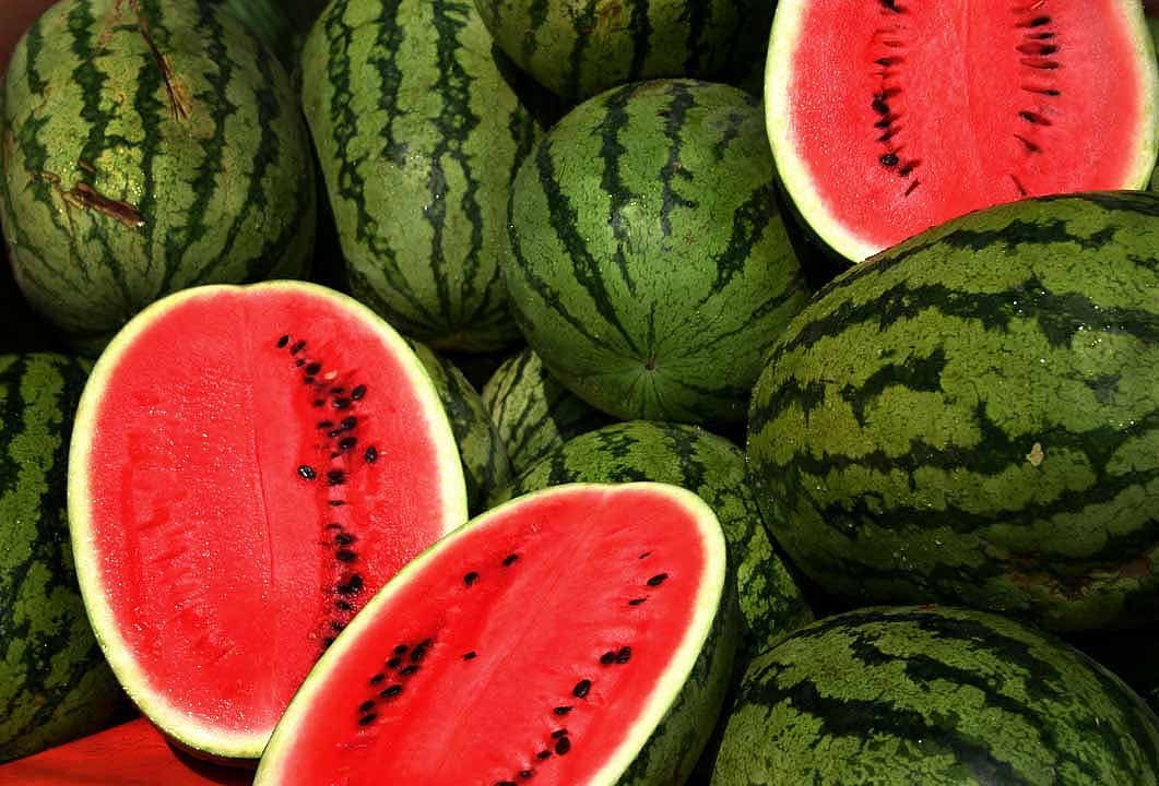 Whole and halved watermelons with green striped rinds and bright red flesh filled with black seeds are piled together, showcasing their fresh and juicy appearance.