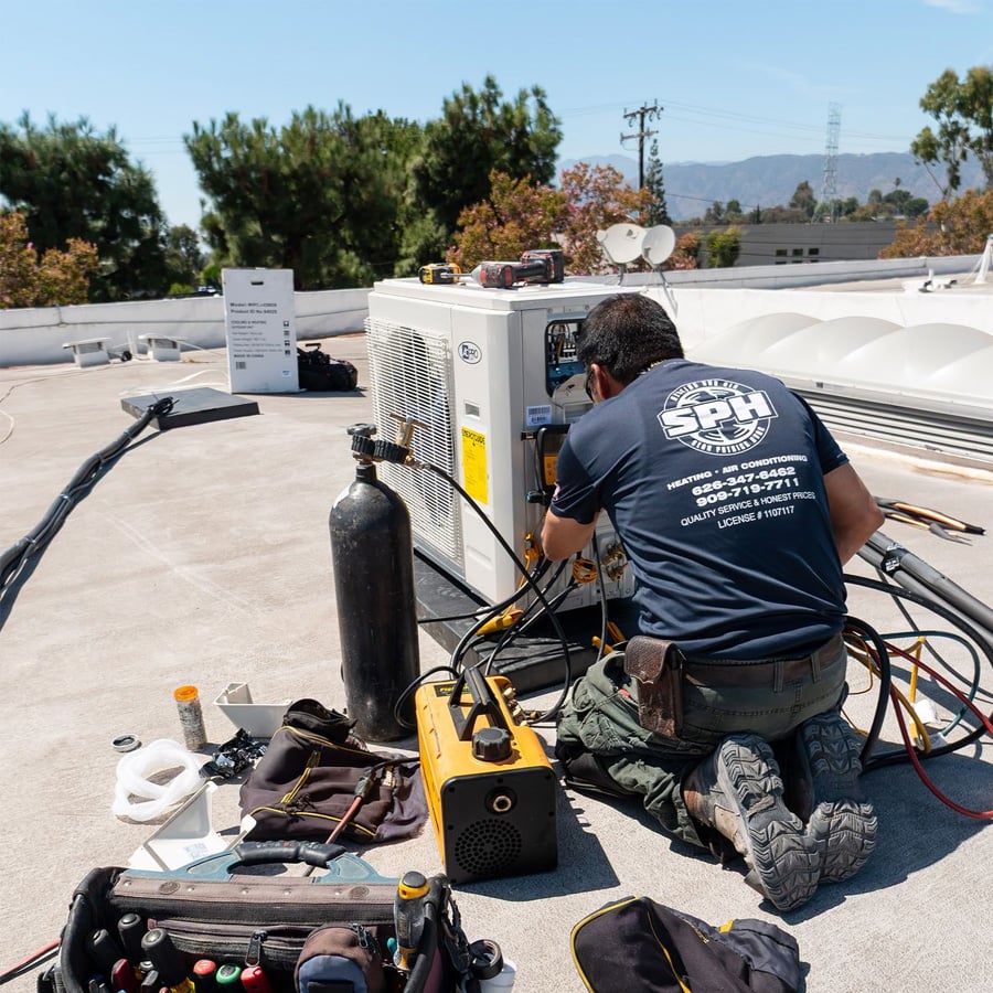 A technician kneels on a rooftop, servicing an HVAC unit with various tools and equipment beside him. Trees and mountains are visible in the background under a clear sky.
