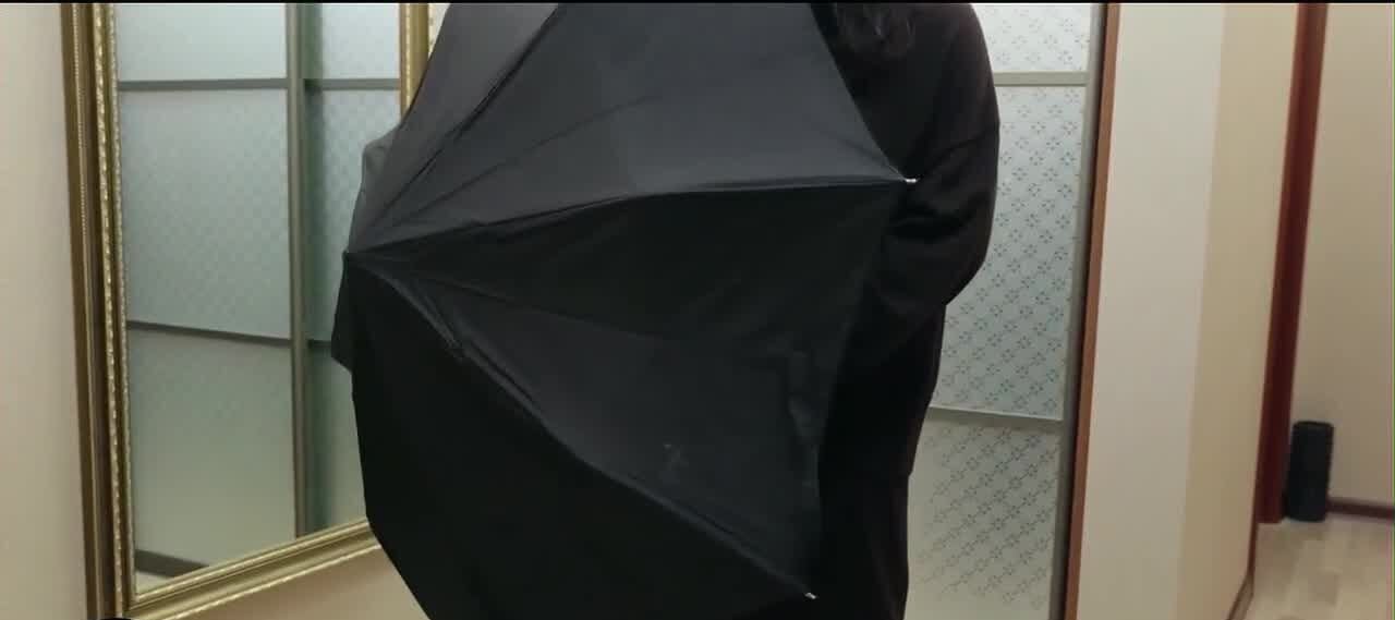 A person dressed in black stands indoors holding an open black umbrella, facing a large mirror with a gold frame. The room has frosted glass sliding doors and light-colored flooring.