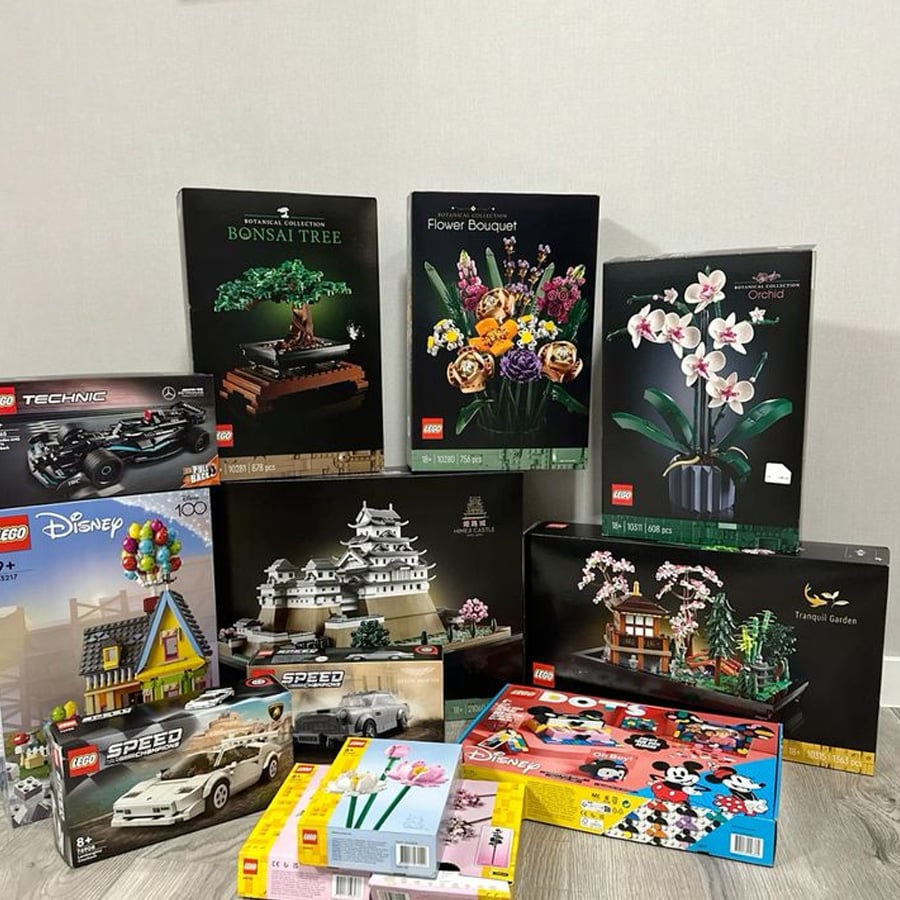 Various LEGO sets in boxes are arranged on a wooden floor against a light-colored wall, including botanical sets, Disney themes, sports cars, and a Japanese castle.