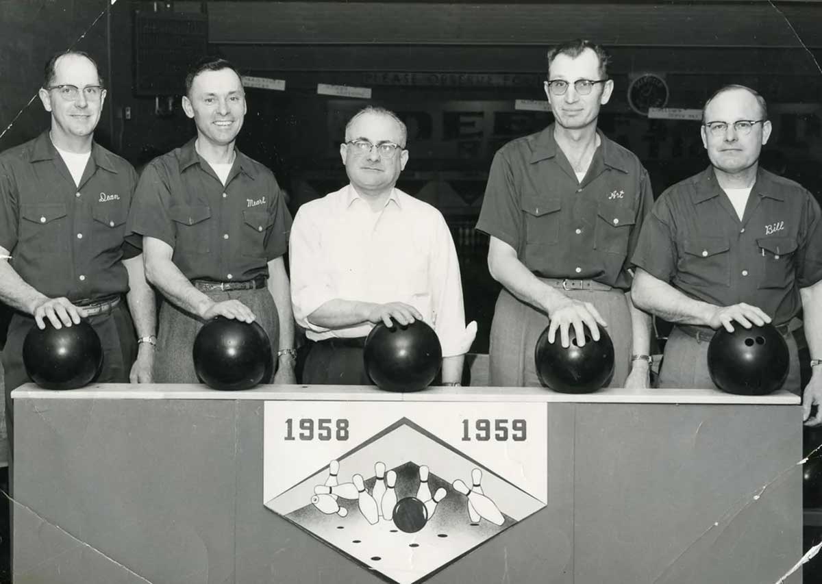 Five men pose behind a sign with bowling illustrations and the years 1958 and 1959; each man rests a hand on a black bowling ball. Four wear matching uniforms and one wears a white shirt.