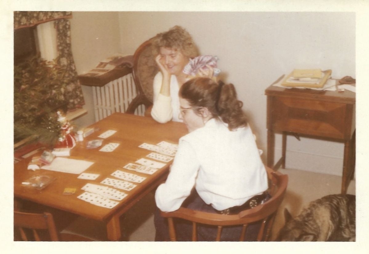 Two women sit at a wooden table playing cards, smiling. There is a small dog in the lower right corner, and papers and books on a side table in the background. The room is softly lit and cozy.