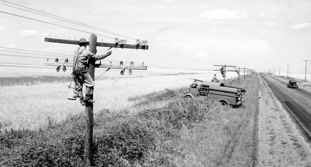 A worker climbs a utility pole to repair power lines near a road in a rural area, while a truck and another worker are visible nearby. Fields stretch into the distance under a partly cloudy sky.