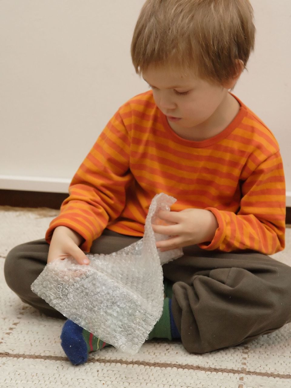 A young child in an orange striped shirt sits on the floor, looking down and playing with a sheet of bubble wrap.