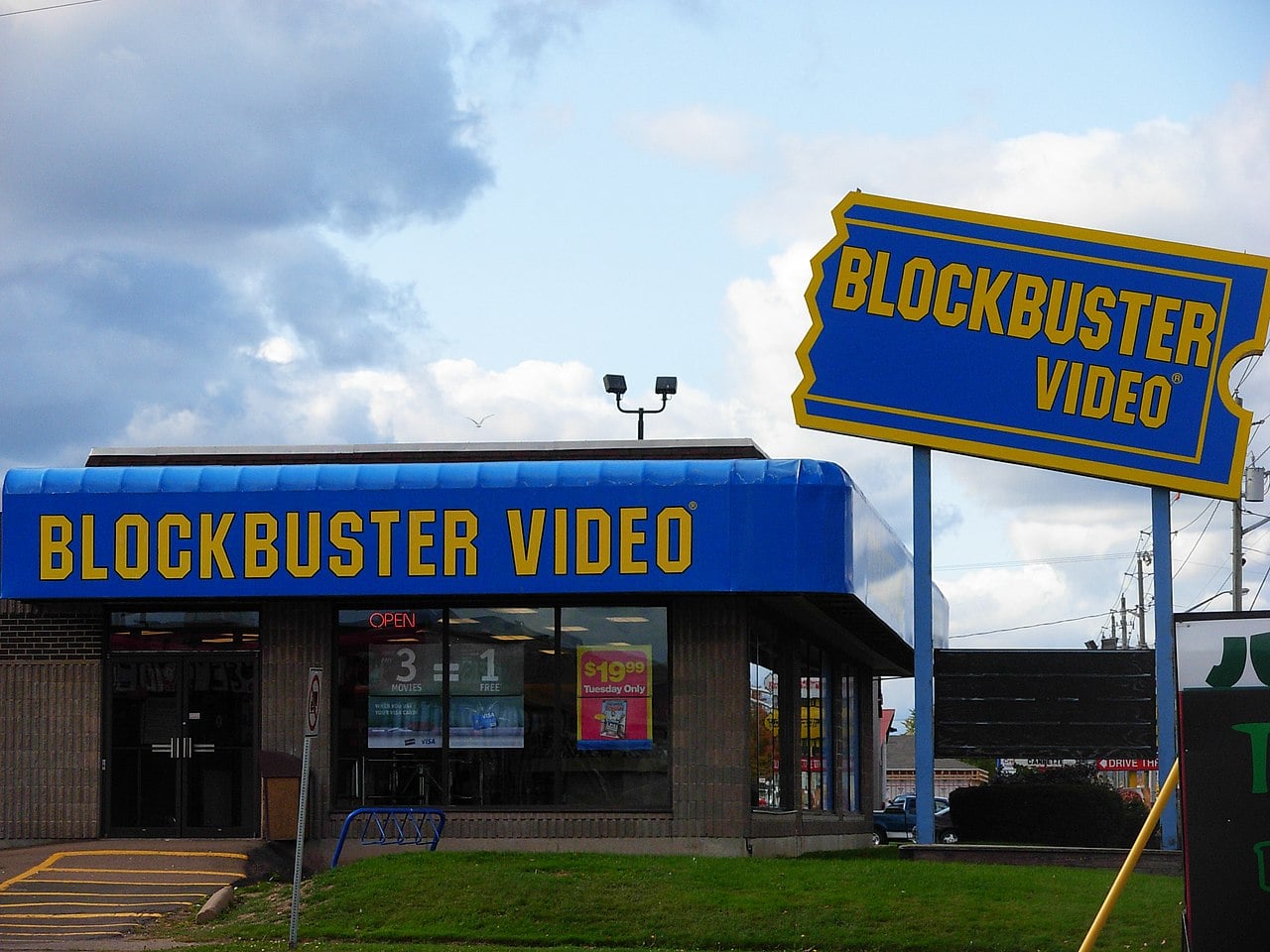A Blockbuster Video store with a large blue and yellow Blockbuster sign outside. The building has a blue awning and windows displaying promotional posters. It appears to be daytime with a cloudy sky.