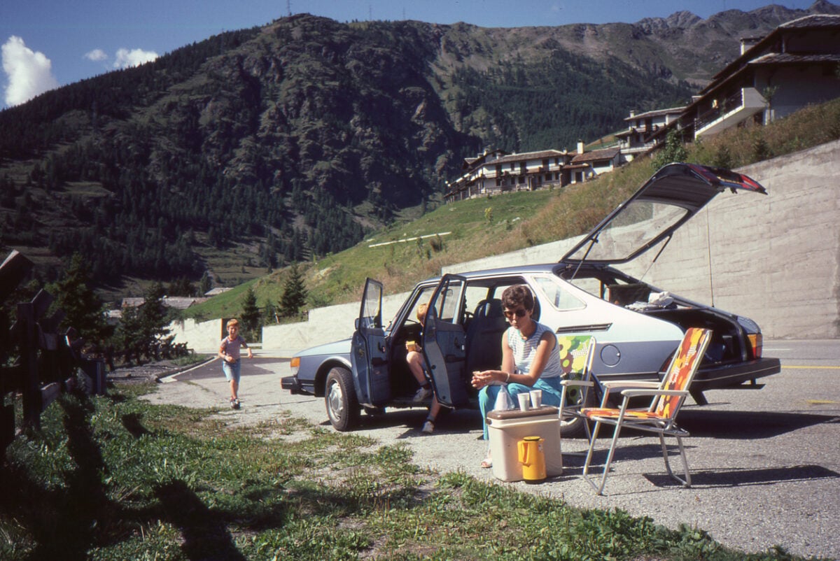 A family picnics by a parked car on a mountain road. A woman sits on a chair, a child stands by the car, and another child walks nearby. The trunk is open. Mountain houses and green hills are visible in the background.