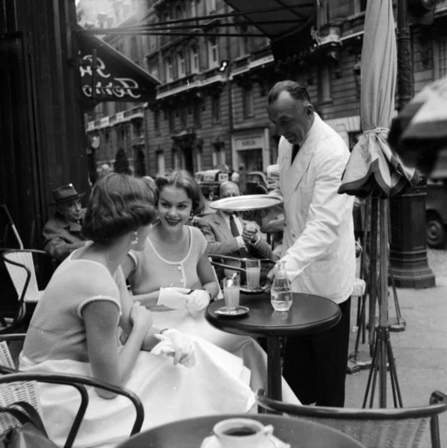 Two women in vintage dresses sit at a café table outdoors, smiling as a waiter in a white jacket serves them drinks. The scene is lively, with people and cars in the background on a city street.