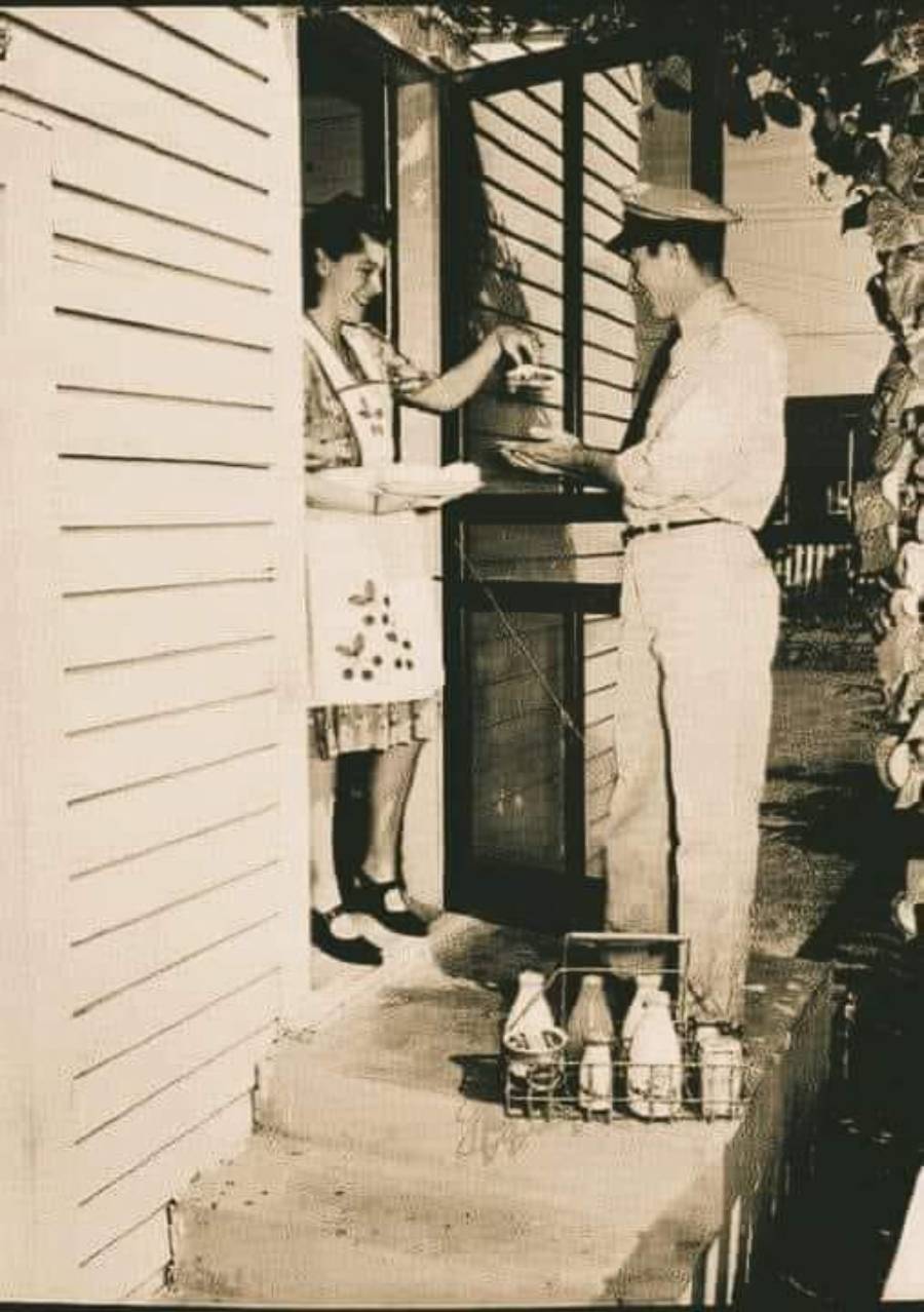 A vintage black-and-white photo of a smiling woman in an apron receiving milk bottles from a milkman at her doorstep. The milkman holds out a bottle while more bottles sit in a carrier on the ground.