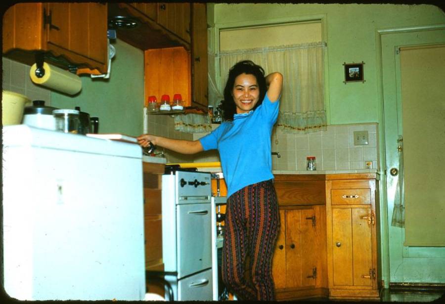A woman in a blue shirt and patterned pants smiles while posing in a retro kitchen with yellow cabinets and a stove.