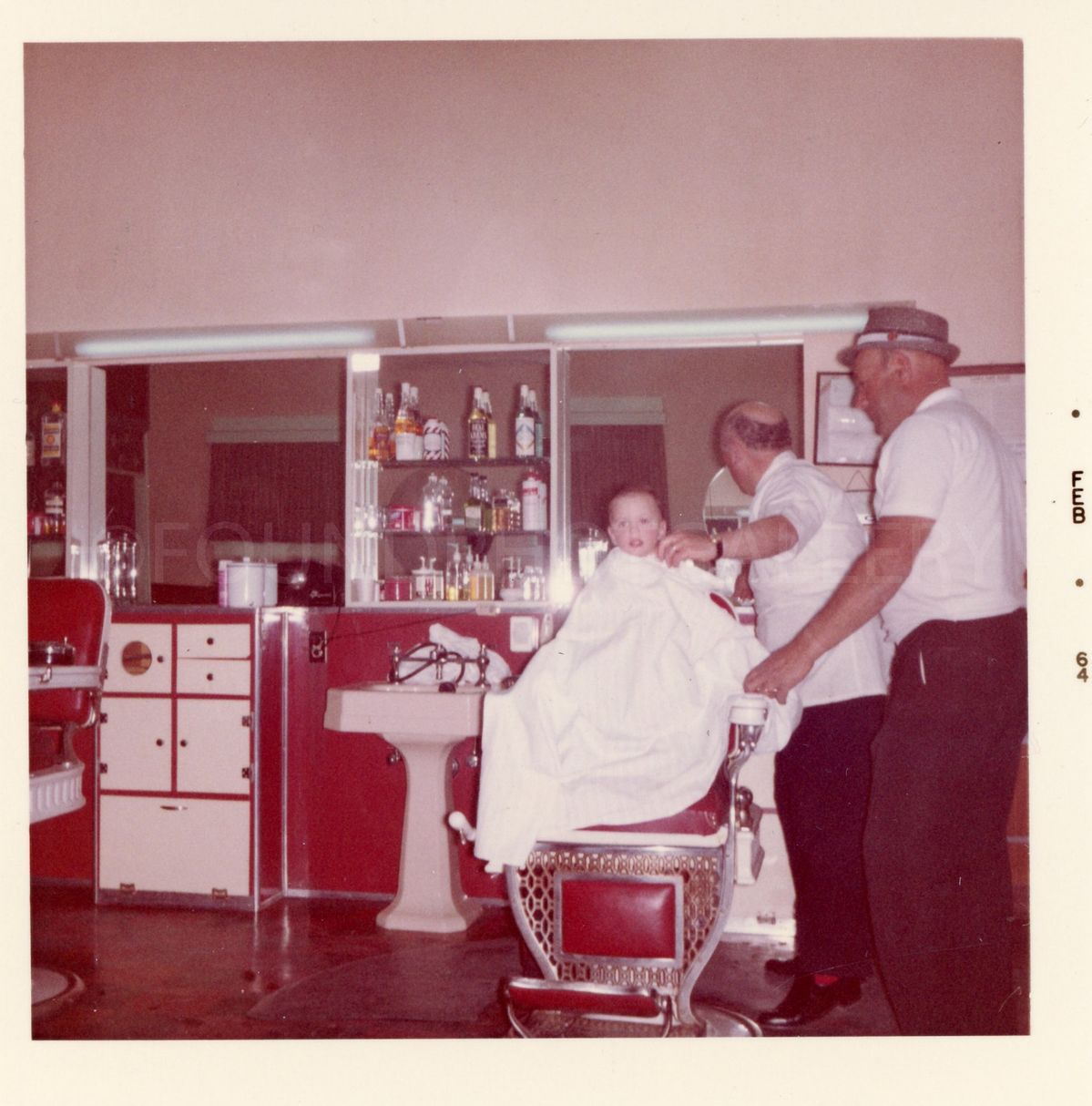A baby sits in a barber chair, covered with a white cape, while two men stand nearby in a vintage barbershop with bottles on shelves behind them.