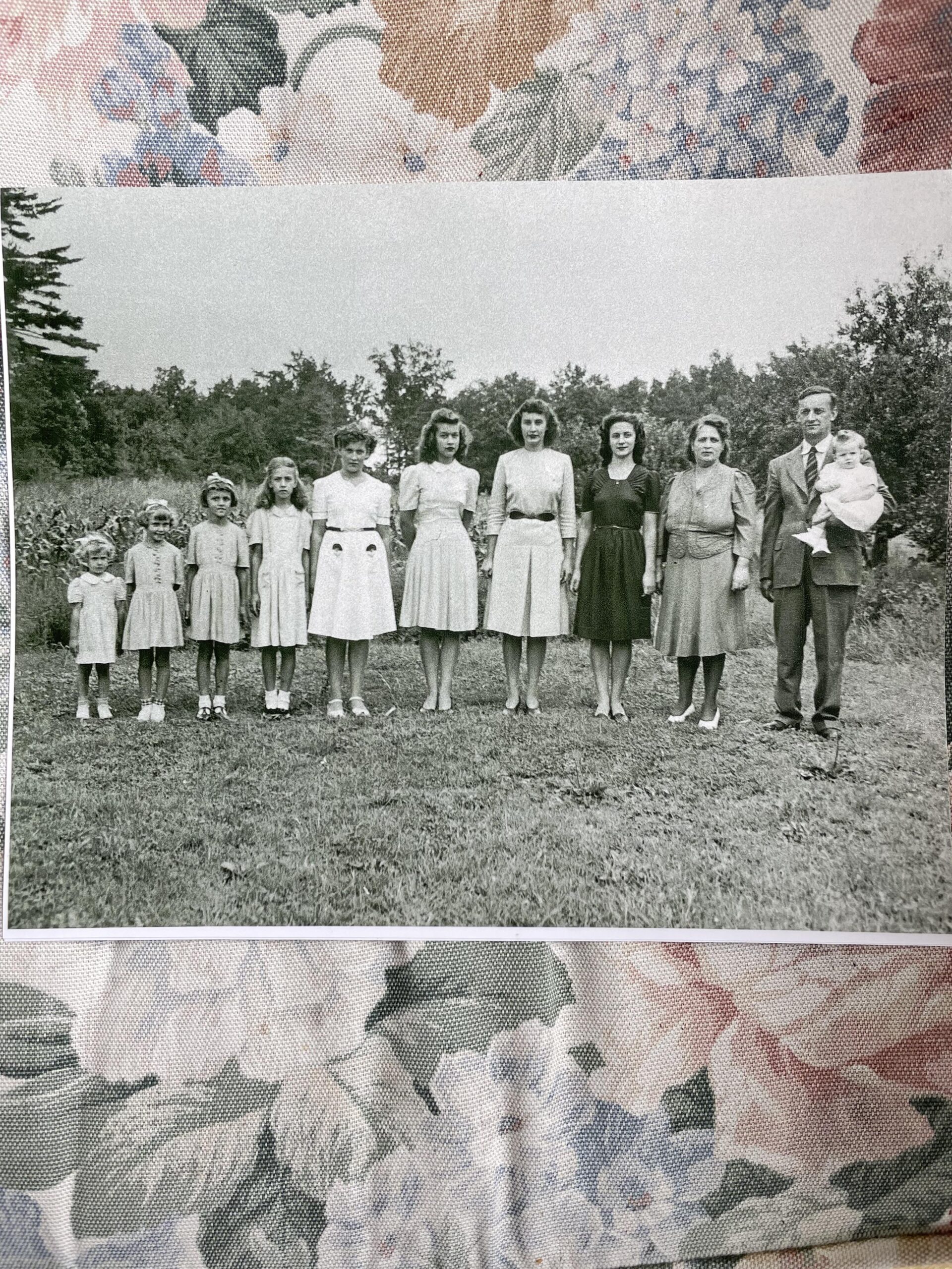 A black-and-white photo of eight girls and one woman standing side by side outdoors, with a man holding a baby on the right. The group is on grass with trees in the background, and the photo rests on a floral-patterned fabric.
