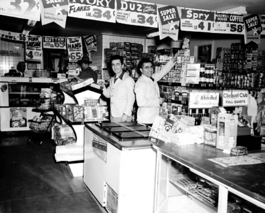Two men in white uniforms stand behind the counter of a vintage grocery store, surrounded by shelves stocked with canned goods and signs advertising sales. The store is bustling with products and promotional signs overhead.