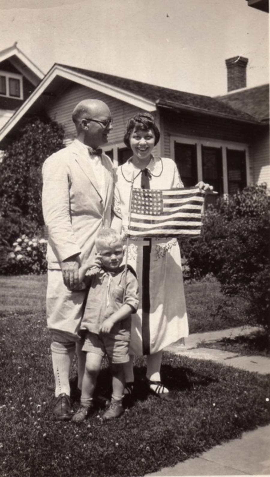A man, woman, and young boy stand outside a house. The woman holds an American flag, and the family smiles. The photo appears to be from the early 20th century.