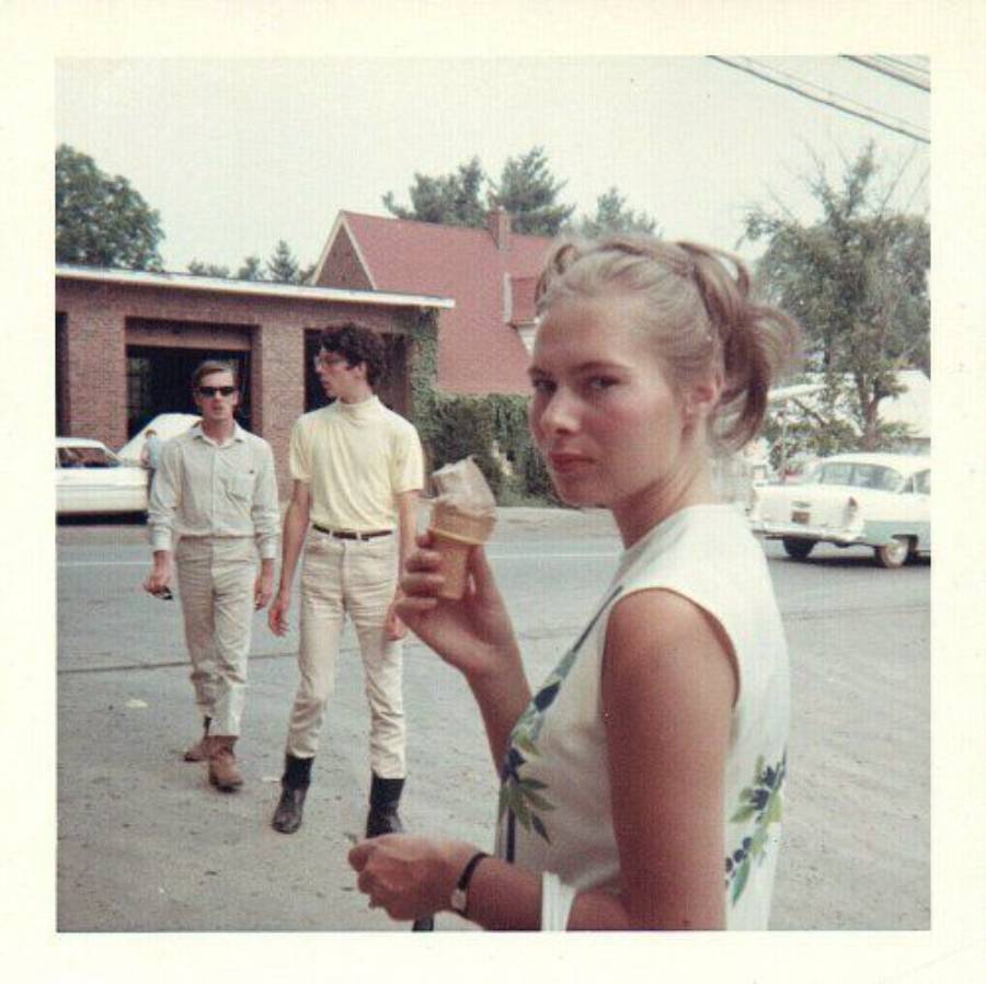 A woman holding an ice cream cone looks at the camera while two men walk behind her on a street lined with cars and trees.