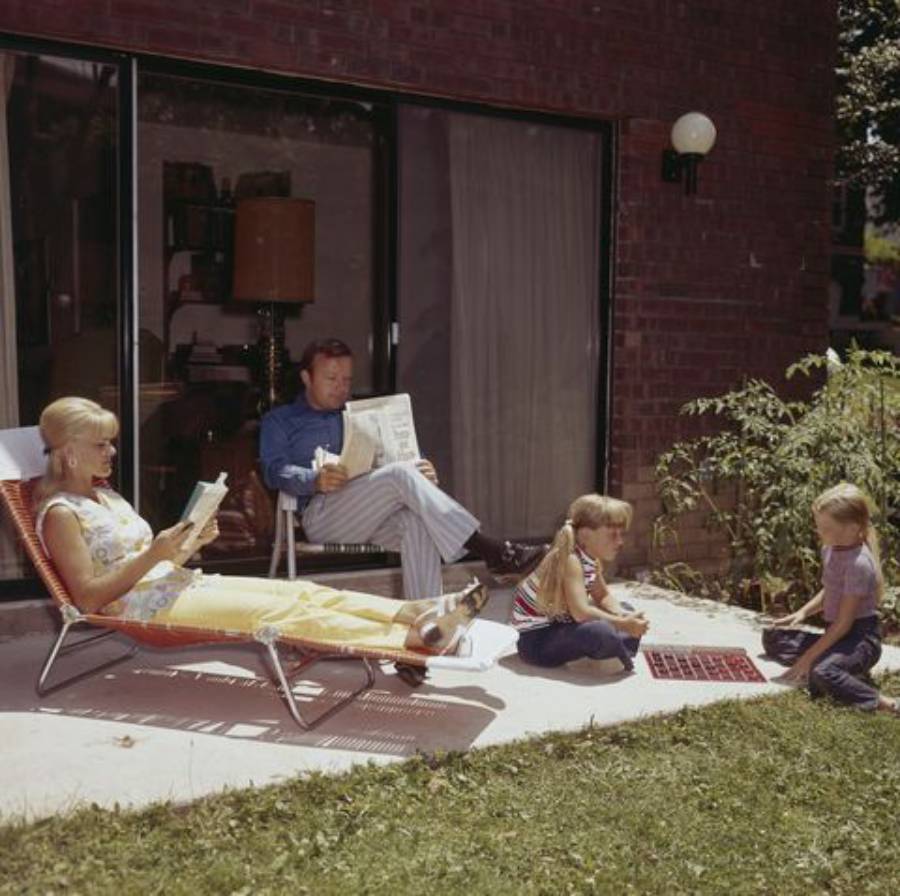 A woman lounges on a chair reading, a man sits nearby reading a newspaper, and two children play a board game on the patio outside a brick house on a sunny day.