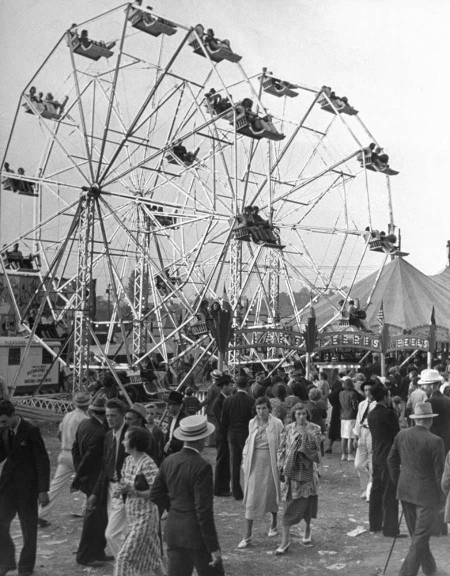 Black-and-white photo of a busy fairground with people walking, two Ferris wheels full of riders, and a circus tent in the background. The crowd is dressed in vintage clothing, suggesting an early 20th-century setting.