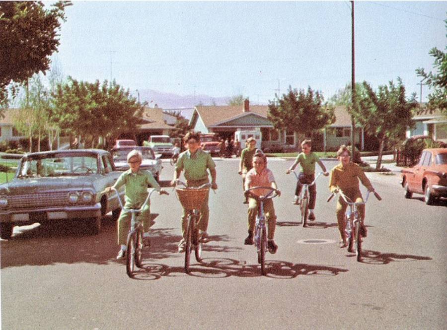 Five children ride bicycles down a suburban street lined with parked cars and single-story houses on a sunny day. Trees and mountains are visible in the background.