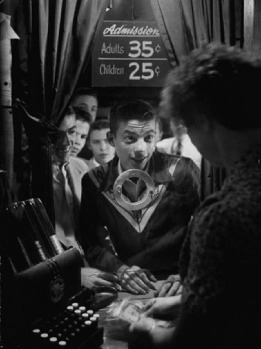 A young man smiles as he buys a ticket at a booth, with several people waiting behind him. A sign above lists admission prices: Adults 35¢, Children 25¢. The ticket seller counts bills at the counter.