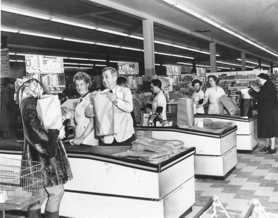 Black and white photo of a busy supermarket checkout area in the past. Cashiers and shoppers, mostly women, interact at the registers with grocery bags, shopping carts, and visible store shelves in the background.