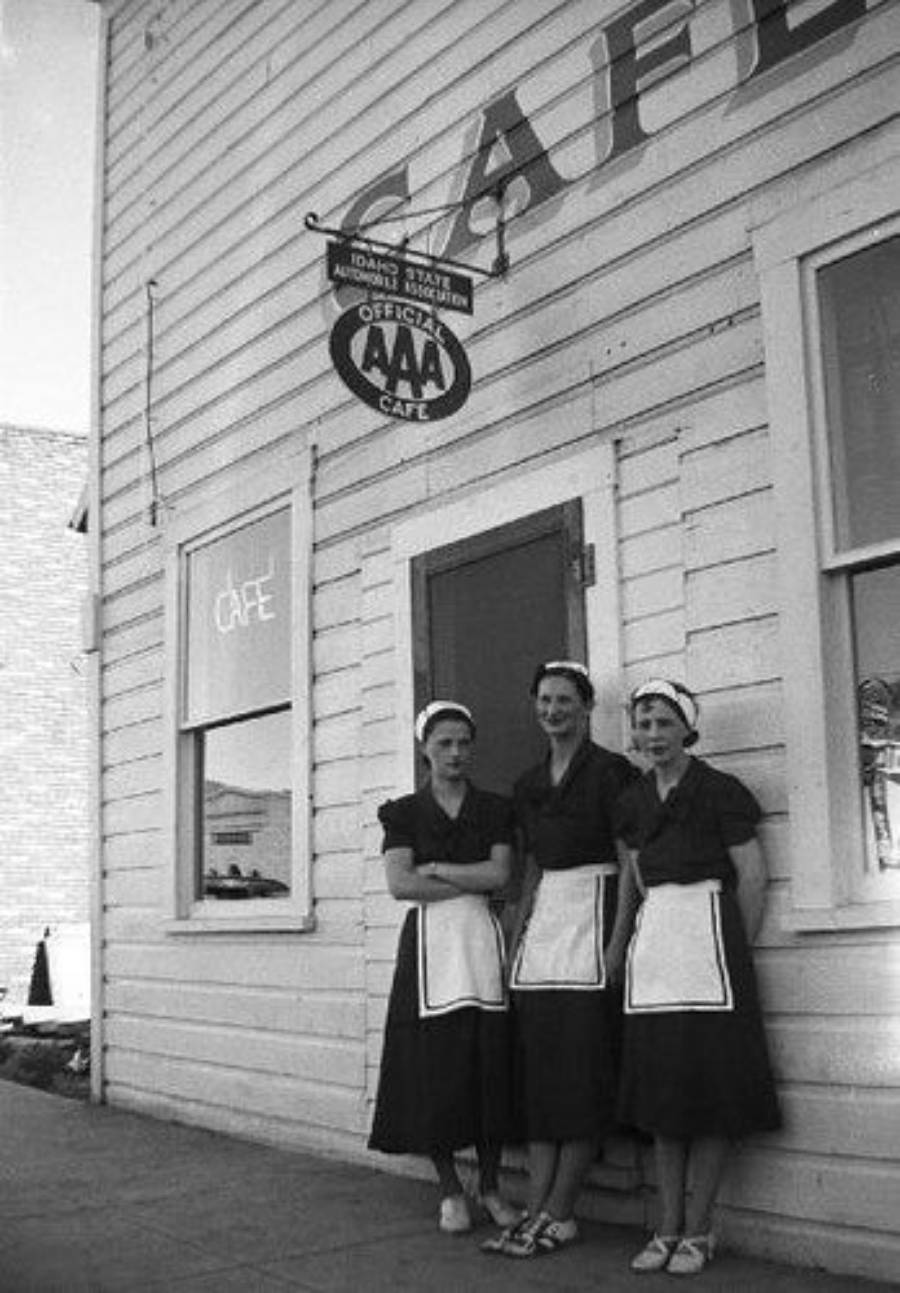 Three women in matching uniforms and aprons stand outside a white wooden cafe building with a neon "CAFÉ" sign and a hanging AAA sign above the door.