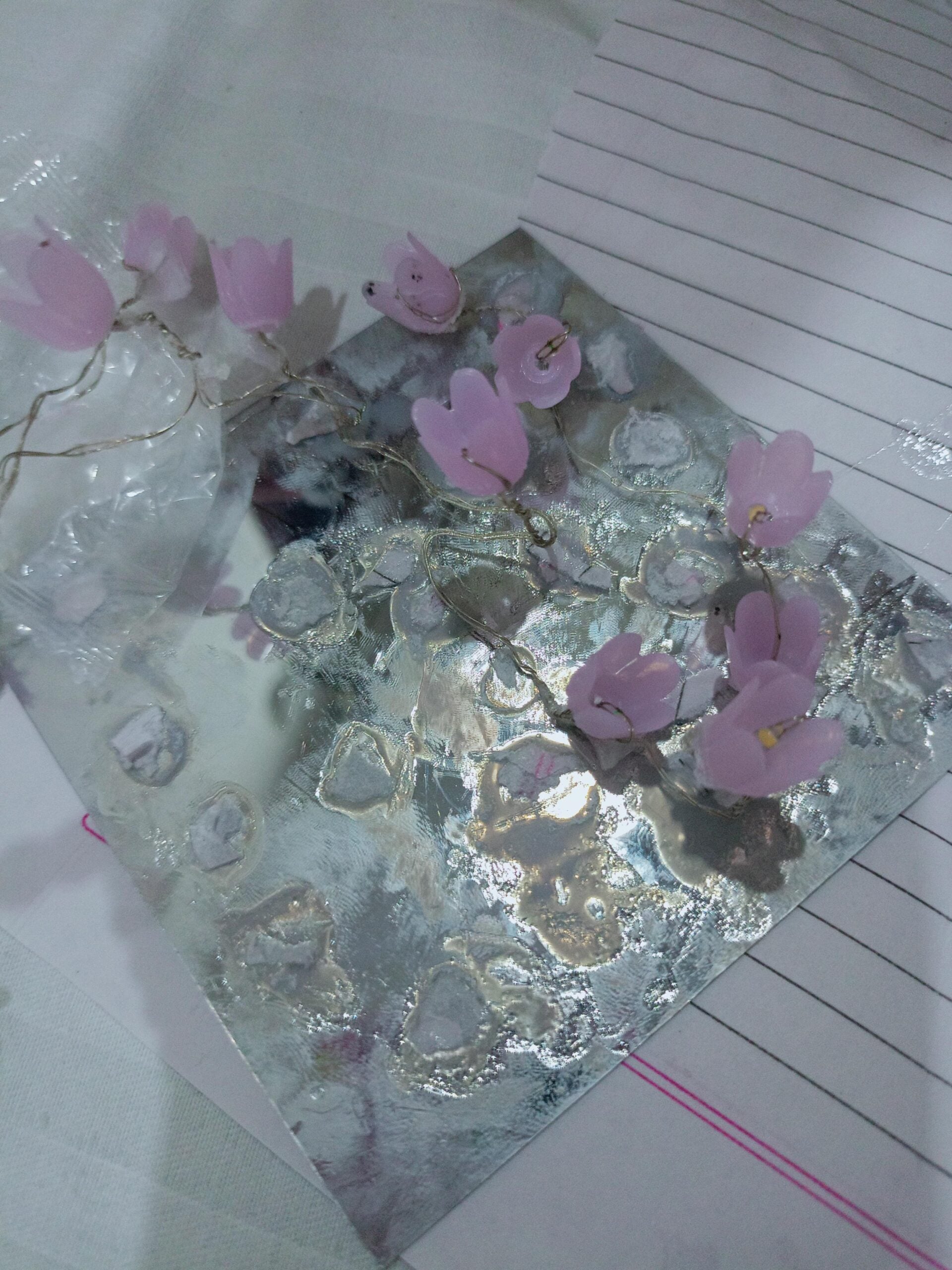 A reflective sheet with silver patches and a pink flower string light is placed on a lined notebook paper on a white surface.