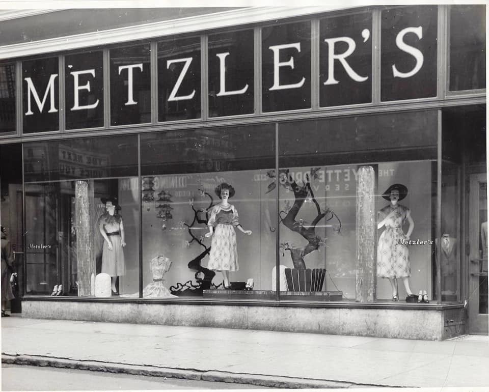 Black-and-white photo of a Metzler’s store window display featuring three mannequins in elegant dresses, surrounded by accessories and decorative elements. Large “METZLER’S” sign is displayed above the window.