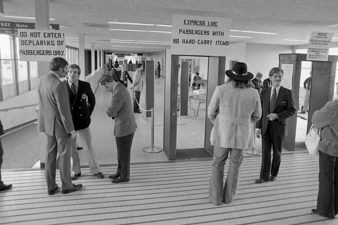 Black and white photo of several men in suits and hats standing in an airport terminal near signs reading "EXPRESS LINE PASSENGERS WITH NO HAND-CARRY ITEMS" and "DO NOT ENTER! DEPLANING OF PASSENGERS ONLY.