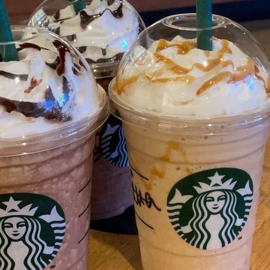 Three Starbucks frappuccinos with whipped cream and caramel or chocolate drizzle on top, served in clear plastic cups with green straws, sitting on a wooden table.