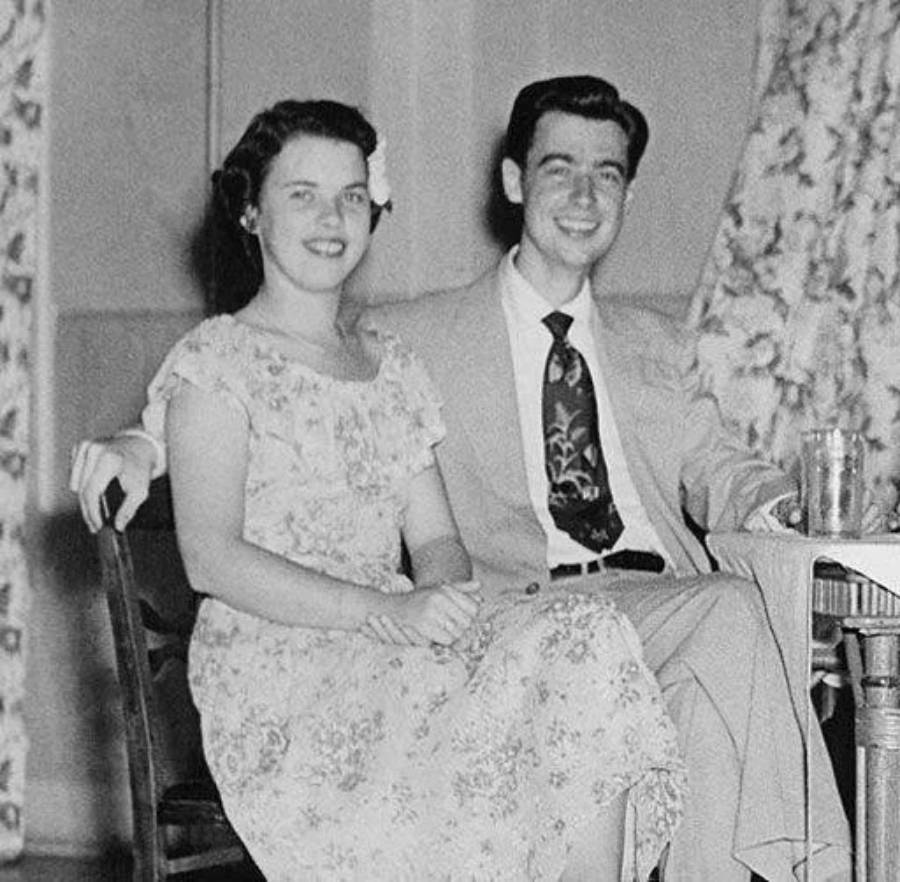 A woman in a floral dress and a man in a suit with a patterned tie sit smiling together at a small table with a glass, in a vintage black and white photo. Curtains and a plain wall are visible in the background.
