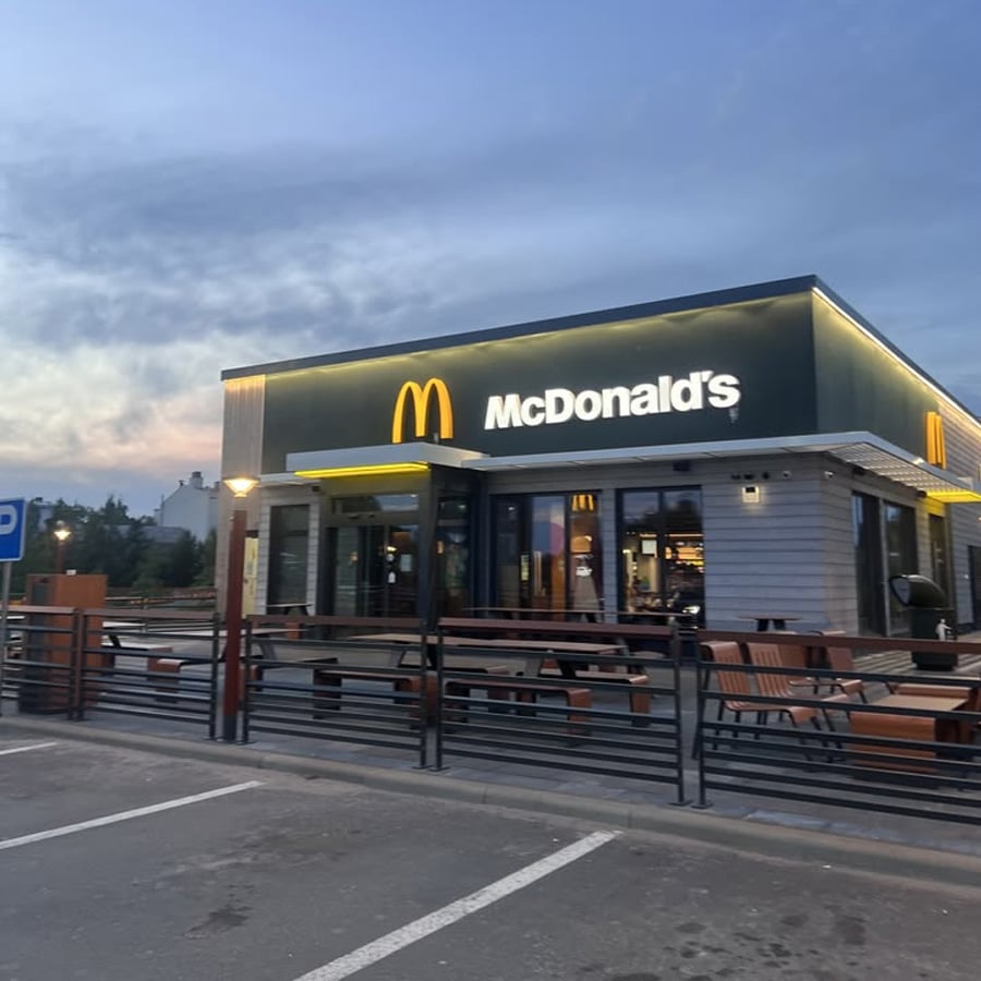 A McDonald’s restaurant with illuminated signs at dusk, showing outdoor seating, empty benches, and a nearby parking area under a cloudy sky.