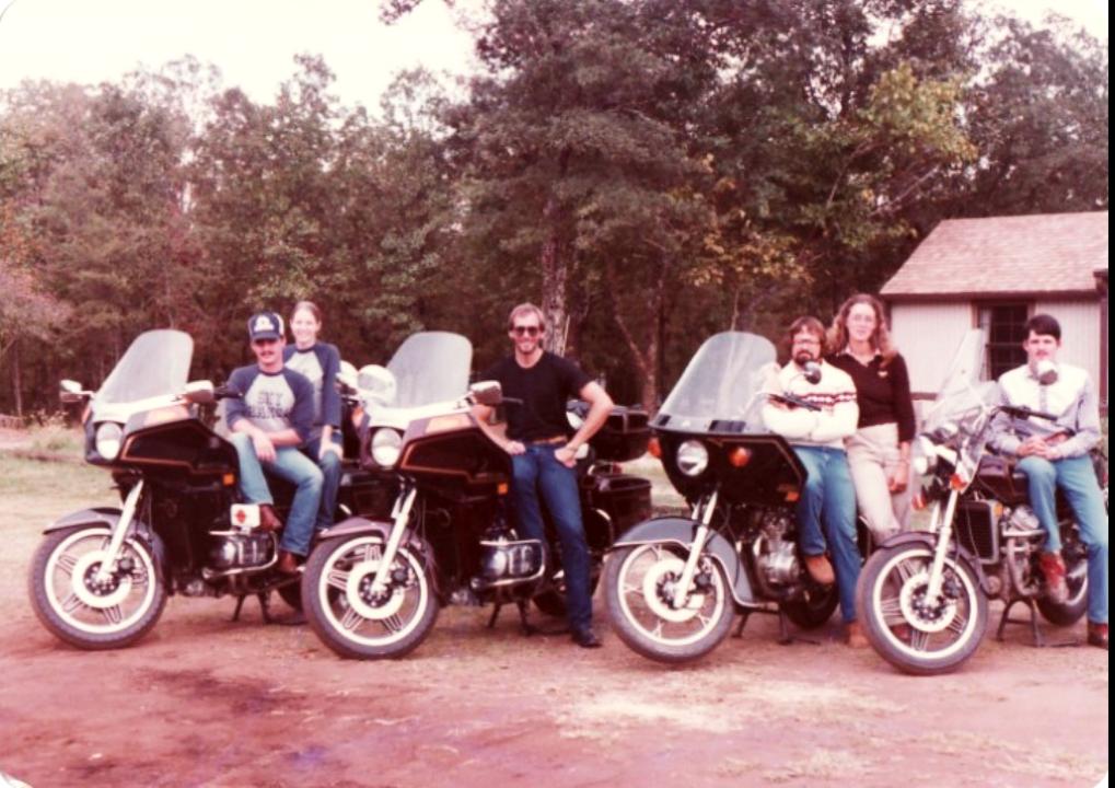 Six people pose outdoors with three motorcycles parked on a dirt surface in front of trees and a small building. Four sit on the bikes, while two stand behind, all smiling at the camera.
