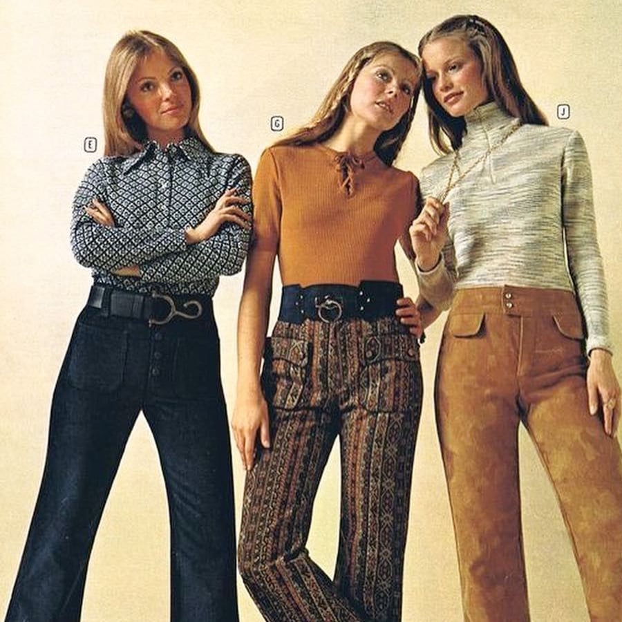 Three women pose in 1970s outfits: one in a patterned blouse and black pants, one in a rust top with patterned pants, and one in a striped turtleneck with tan suede pants, all with long hair.