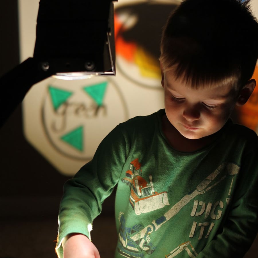 A young boy stands by an overhead projector, illuminated by its light. Colorful shapes and letters are projected onto the wall behind him. He is focused on something below the projector, wearing a green shirt with construction vehicles.