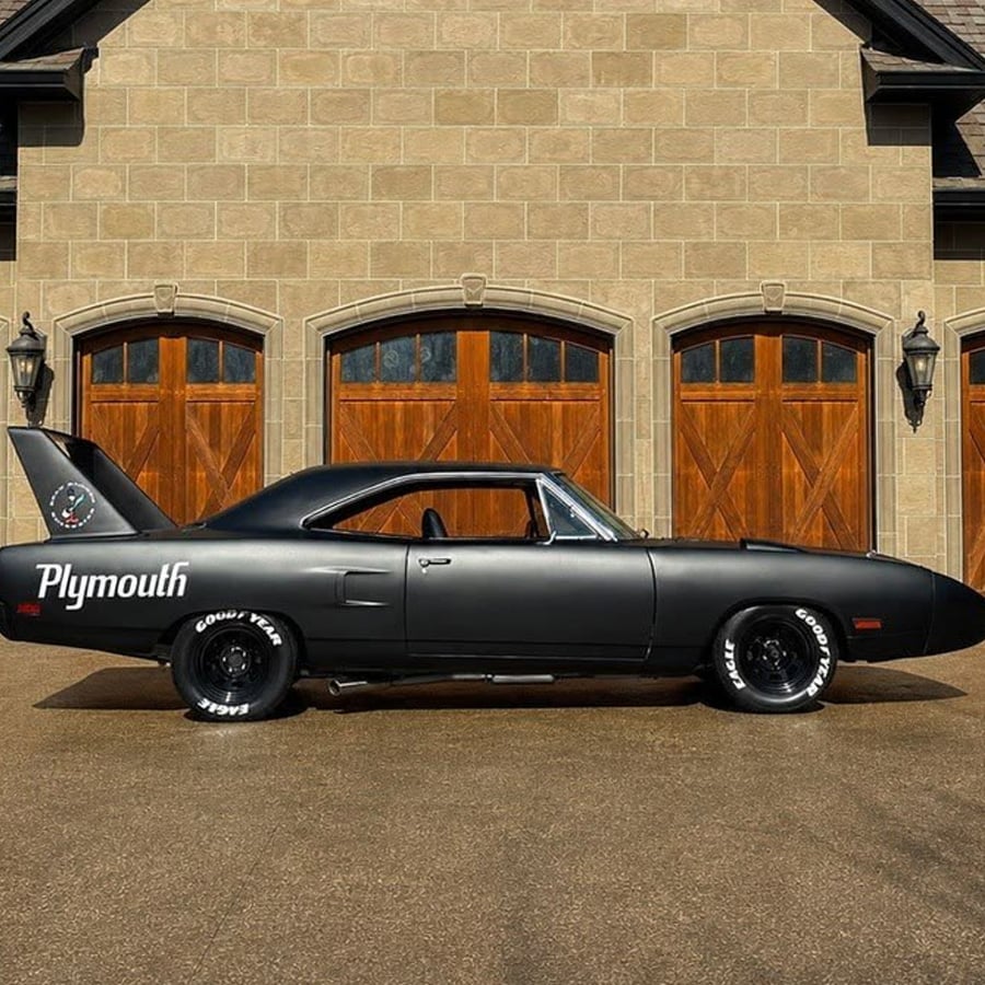 A matte black Plymouth Superbird muscle car with a large rear wing is parked in front of a stone house with three wooden garage doors.