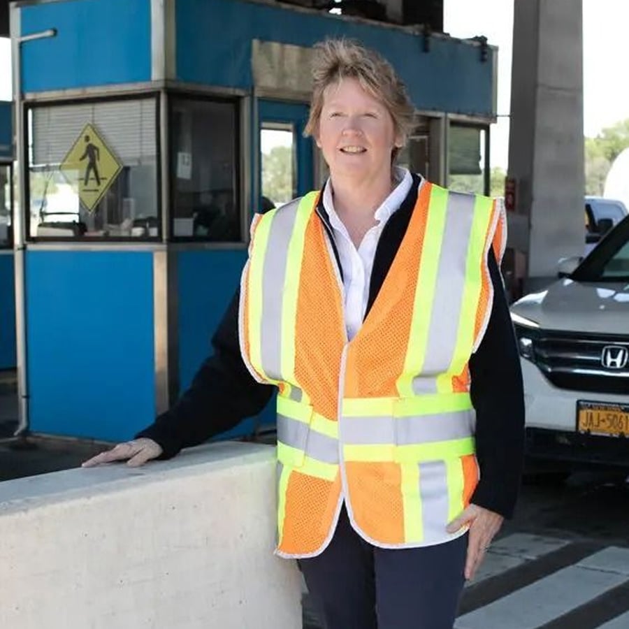 A woman wearing a reflective safety vest stands and smiles near a toll booth and a white vehicle. A pedestrian crossing sign is visible in the background.