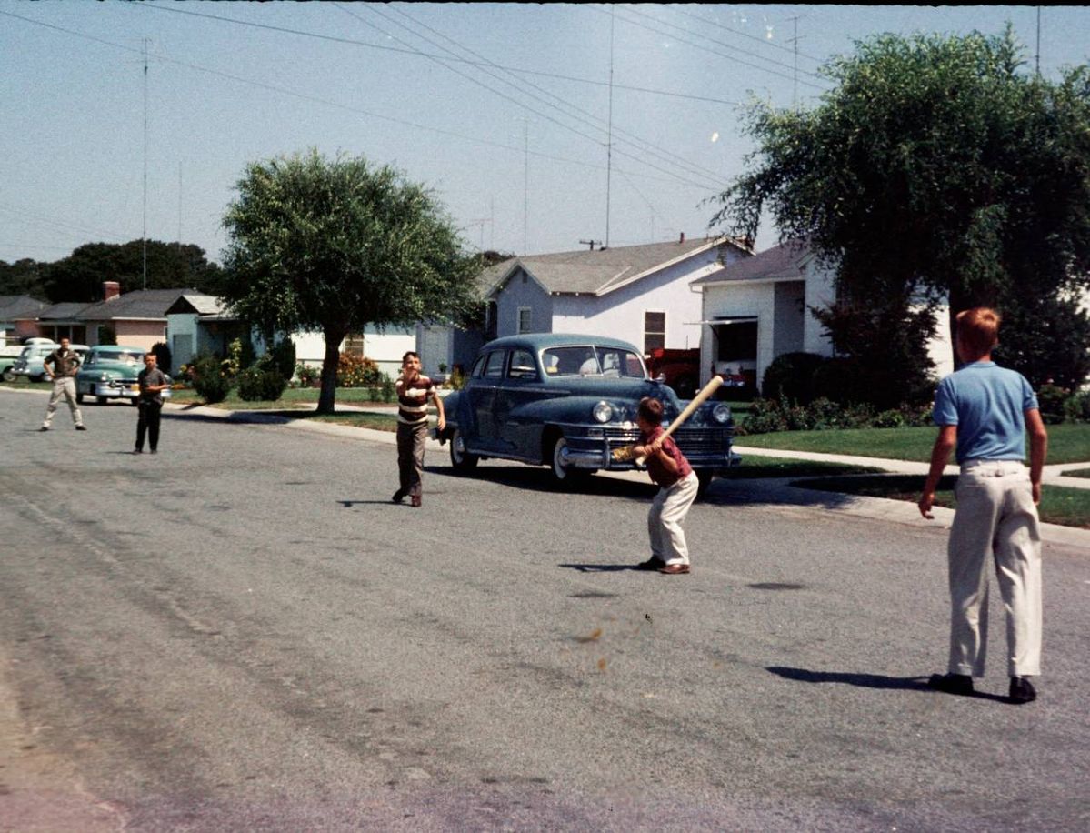 Children play a game of baseball on a suburban street lined with parked cars and houses, while trees and lawns frame the scene on a sunny day.