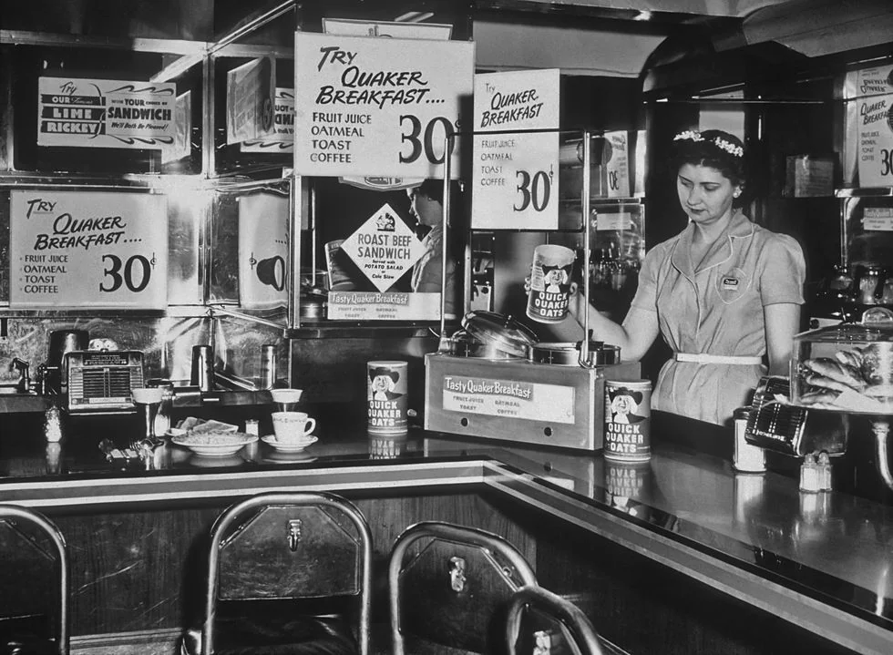 A waitress stands behind a 1950s diner counter with Quaker Oats cans and breakfast signs. The counter displays food and beverages, and mirrored walls reflect the retro decor and advertisements.