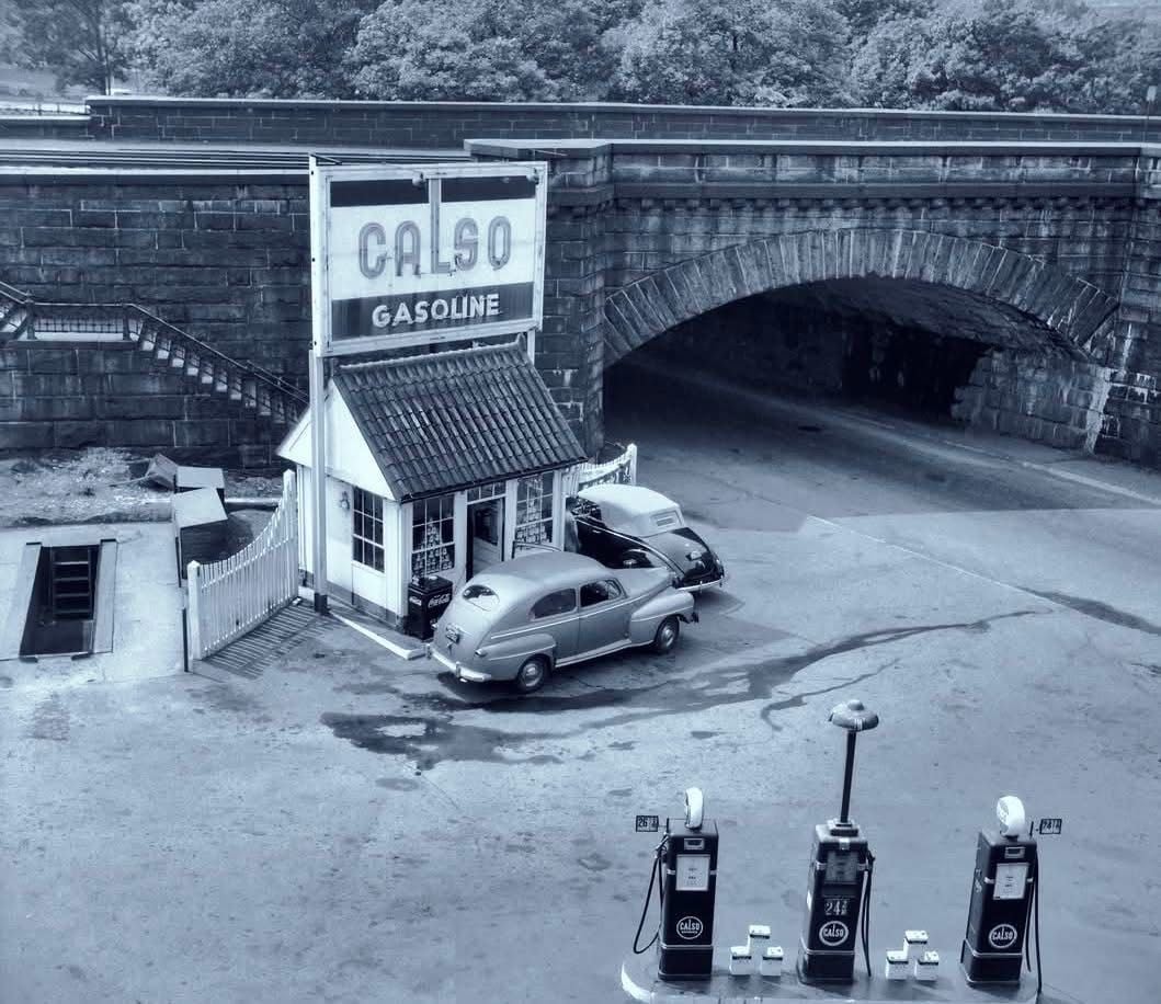 A vintage gas station labeled "CALSO GASOLINE" sits beside an arched stone bridge. Two classic cars are parked nearby, and three old-fashioned gas pumps are visible in the foreground.